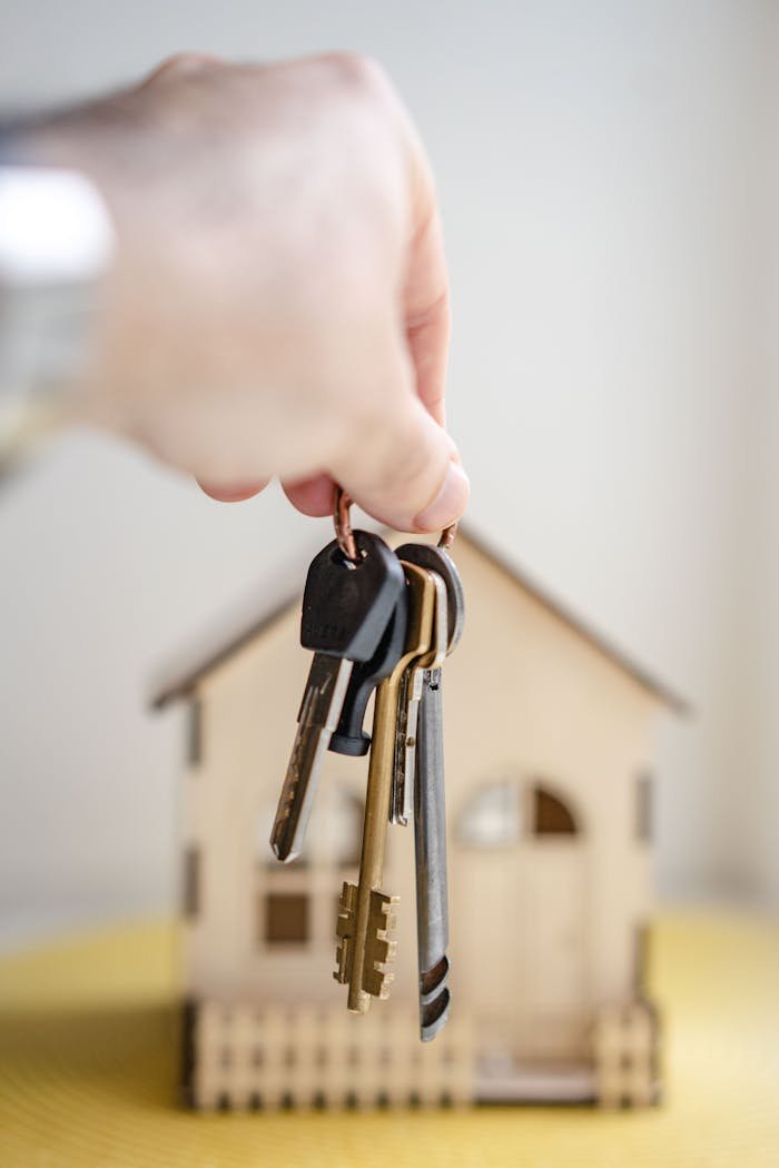 about-us Close-up of a hand holding keys with a miniature wooden house in the background, symbolizing real estate investment.